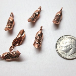May include: Five copper-colored metal charms shaped like miniature clogs, each featuring a stamped flower design on the toe and a small loop at the heel. A United States dime is positioned next to the charms to provide a sense of scale.