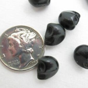 May include: A close-up of six black skull beads, with one bead resting on a US dime for size comparison. The beads are smooth and glossy.