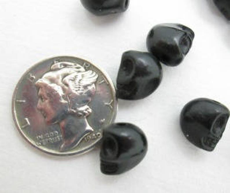 May include: A close-up of six black skull beads, with one bead resting on a US dime for size comparison. The beads are smooth and glossy.