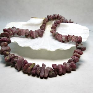 May include: A strand of pink ruby gemstone chips strung on a wire. The chips are irregular in shape and size, and have a natural, rough texture. The strand is displayed on a white seashell.