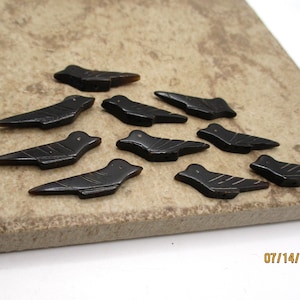 May include: A collection of ten small, dark brown bird-shaped beads. Each bead features a smooth, polished surface with carved lines representing wings and a tail. The bird figures are arranged in a scattered formation, showcasing their uniform shape and dark, glossy finish.