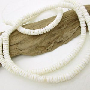 May include: White shell necklace with a layered design. The necklace is composed of small, flat, rounded shell pieces strung together. The necklace is displayed on a piece of driftwood.
