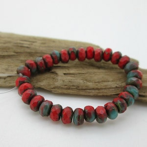 May include: A strand of faceted beads in a vibrant red hue, accented with patches of teal and dark green. The beads are strung together, creating a circular shape. The beads are resting on a piece of driftwood.