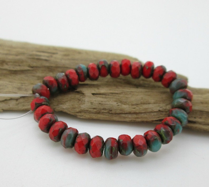 May include: A strand of faceted beads in a vibrant red hue, accented with patches of teal and dark green. The beads are strung together, creating a circular shape. The beads are resting on a piece of driftwood.