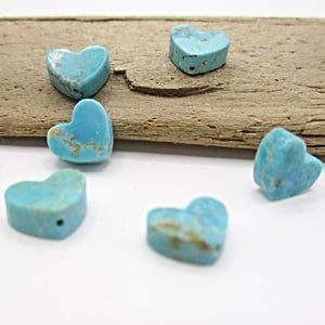 May include: Several turquoise heart-shaped beads with natural variations in color and texture. The beads are scattered on a white surface with a piece of driftwood in the background. These beads are suitable for jewelry making.
