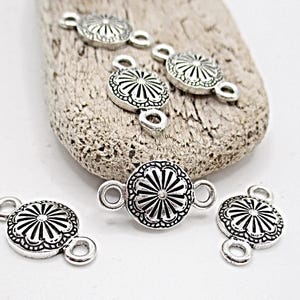 May include: Silver-toned metal jewelry connectors with a floral design. The connectors have a circular shape with a detailed pattern and loops on each end for attachment. Several connectors are arranged on a light-colored, textured surface.