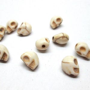 May include: A collection of small, off-white skull beads with brown detailing. The beads are scattered across a white surface, showcasing their carved features, including eye sockets, nasal cavity, and jawline. These beads are ideal for jewelry making.