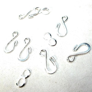 May include: A collection of silver-toned metal S-hooks and figure-eight shaped jewelry findings. These small, curved components feature smooth, reflective surfaces and open loops, designed for connecting jewelry pieces or other small craft items.