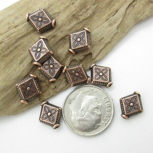 May include: A group of ten copper-colored metal beads with a square shape and a floral design. The beads are scattered on a wooden surface.