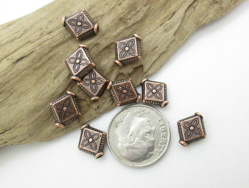 May include: A group of ten copper-colored metal beads with a square shape and a floral design. The beads are scattered on a wooden surface.