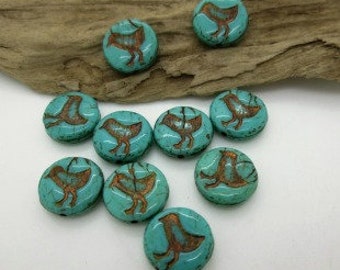 May include: A collection of turquoise-colored, round beads with a bird design in a copper color. The beads are scattered on a piece of driftwood, showcasing their detailed avian artwork.