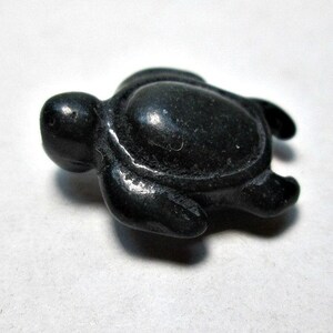 May include: A small, black turtle figurine. The turtle is carved from a dark, possibly stone-like material. It has a rounded shell, a small head, and four flipper-like appendages. The figurine is set against a white background.