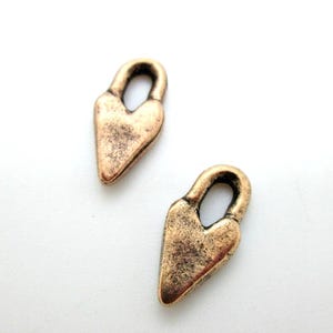 May include: Two antique bronze-colored heart-shaped charms with a loop at the top. The charms have a textured, aged appearance, suggesting a vintage or handcrafted aesthetic. The background is plain white.