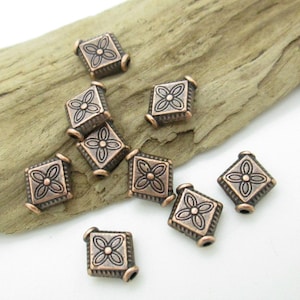 May include: Ten copper-colored square beads with a raised floral design. The beads have a hole through the center for stringing.