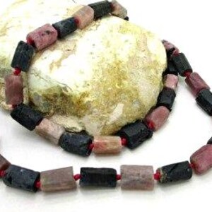 May include: A necklace featuring rectangular beads in shades of pink, black, and brown. The beads are strung together with red string. The necklace is displayed on a light-colored, textured surface.