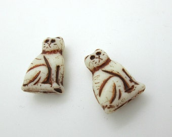 May include: Two white ceramic cat beads with brown details. The cats are sitting with their tails curled around their legs.