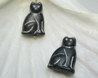 May include: Two black cat shaped glass beads with silver accents. The beads are sitting on a white seashell.