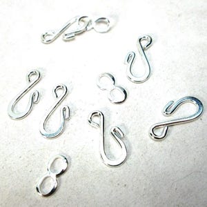 May include: A collection of silver-colored jewelry clasps and connectors. The clasps are in various shapes, including S-hooks and figure-eight designs, suitable for necklaces and bracelets. The pieces are displayed against a light background.