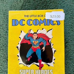 May include: A yellow book with the title "The Little Box of DC Comics: Super Heroes" featuring a comic book illustration of Superman in a red and blue suit flying through a torn piece of paper. The book also includes the text "Batman Superman Wonder Woman" and "TASCHEN".