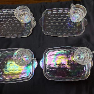 May include: A set of four rectangular glass snack trays with integrated handles and matching glass cups. Each piece features a textured, grid-like pattern and an iridescent finish that reflects light in shades of pink, green, and blue.