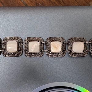 May include: A silver-toned bracelet featuring seven square, off-white, inlaid stones. Each stone is set within a decorative, square frame. The bracelet has a toggle clasp and is designed for wrist wear.