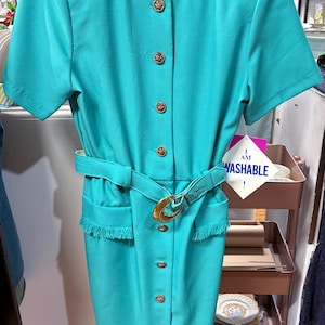 May include: A short-sleeved teal dress featuring a round neckline, a front button closure with gold-toned buttons, and a matching belt with a gold-toned buckle. The dress includes two front pockets with fringe trim and a tag that reads I AM WASHABLE.