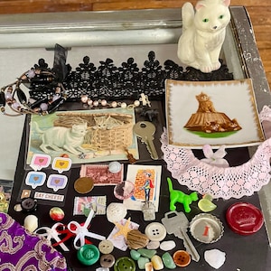 May include: A collection of vintage and modern trinkets, including a porcelain cat figurine, a small plate with a cat design, a postcard with a cat image, a green plastic cat toy, a variety of buttons, beads, and other small objects.  The items are arranged on a black surface with a lace doily and a purple velvet pouch.