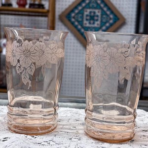 May include: Four vintage pink-tinted drinking glasses. Each glass features a floral design etched near the top. The glasses have a slightly flared rim and a ribbed base. The glasses are arranged on a white lace tablecloth.