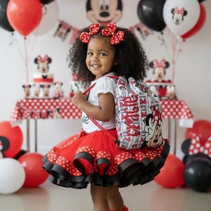 K&ouml;nnte beinhalten: Ein junges M&auml;dchen in einem rot-schwarzen Polka-Dot-Tutu, einem wei&szlig;en Hemd und schwarzen Turnschuhen mit roten Akzenten. Sie tr&auml;gt einen Rucksack und Haarschleifen im Minnie-Maus-Design. Ballons und eine Torte mit Minnie-Maus-Dekorationen sind im Hintergrund.