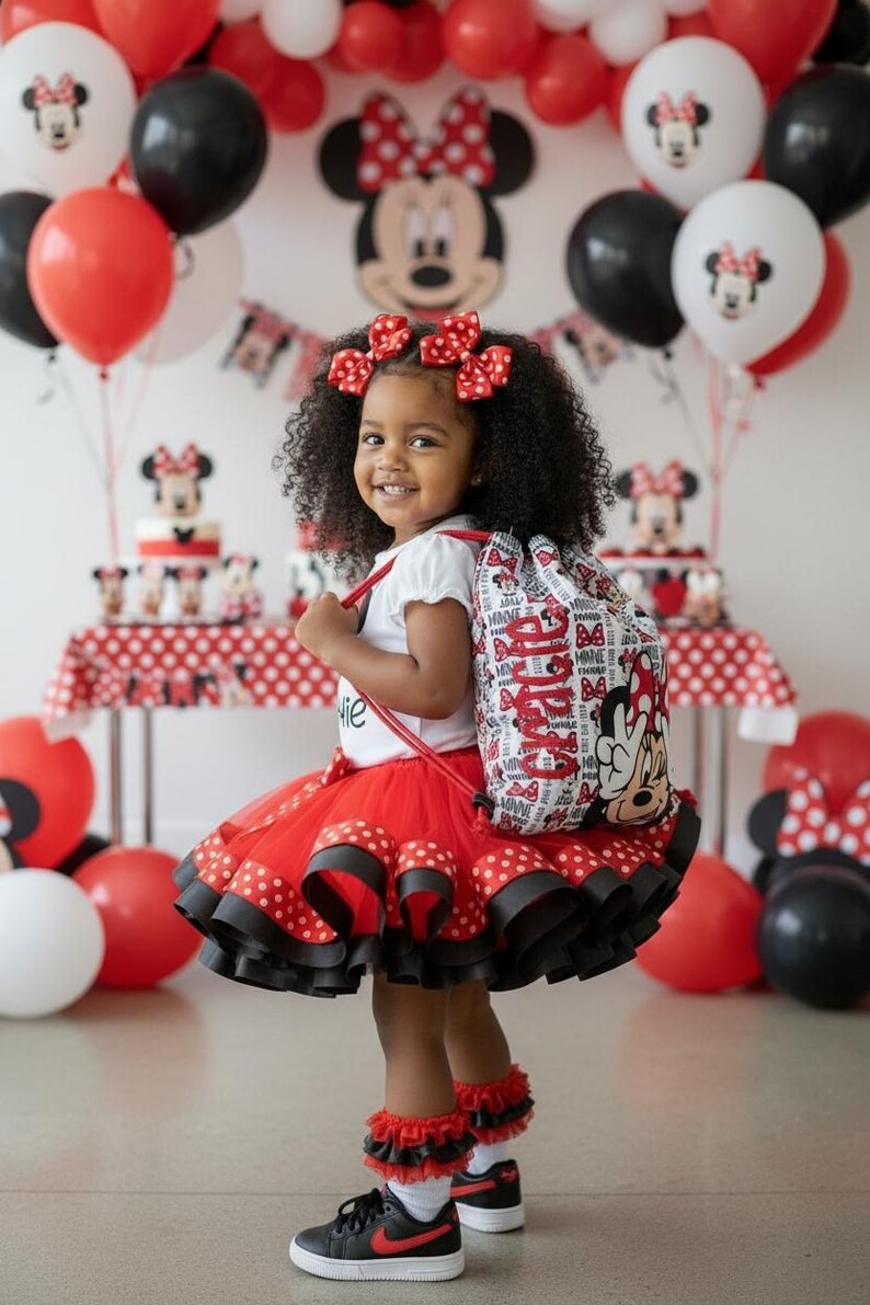 K&ouml;nnte beinhalten: Ein junges M&auml;dchen in einem rot-schwarzen Polka-Dot-Tutu, einem wei&szlig;en Hemd und schwarzen Turnschuhen mit roten Akzenten. Sie tr&auml;gt einen Rucksack und Haarschleifen im Minnie-Maus-Design. Ballons und eine Torte mit Minnie-Maus-Dekorationen sind im Hintergrund.