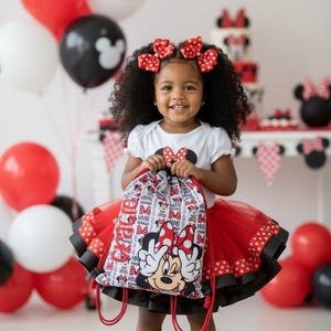 K&ouml;nnte beinhalten: Ein junges Kind l&auml;chelt und h&auml;lt einen Minnie-Maus-Kordelzugbeutel. Sie tr&auml;gt ein wei&szlig;es Oberteil mit einer Minnie-Maus-Schleife, einen roten T&uuml;llrock und schwarze Turnschuhe. Rote und schwarze Luftballons und eine Minnie-Maus-Torte sind im Hintergrund.