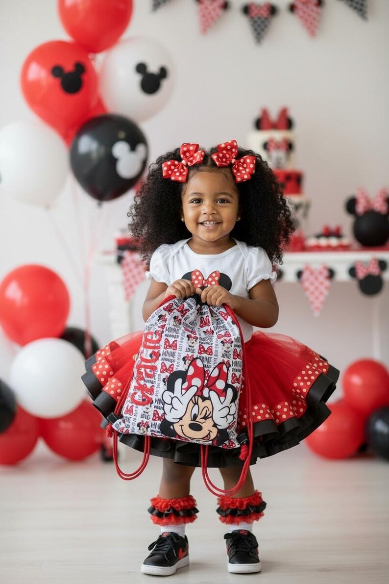 K&ouml;nnte beinhalten: Ein junges Kind l&auml;chelt und h&auml;lt einen Minnie-Maus-Kordelzugbeutel. Sie tr&auml;gt ein wei&szlig;es Oberteil mit einer Minnie-Maus-Schleife, einen roten T&uuml;llrock und schwarze Turnschuhe. Rote und schwarze Luftballons und eine Minnie-Maus-Torte sind im Hintergrund.