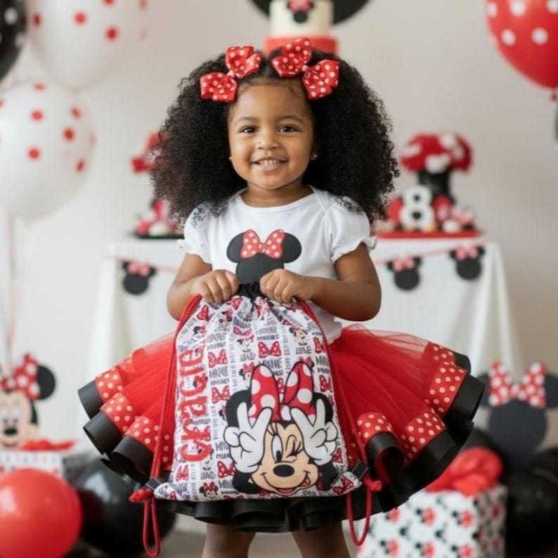 K&ouml;nnte beinhalten: Ein junges Kind tr&auml;gt ein wei&szlig;es T-Shirt mit einem Minnie-Maus-Motiv, einen roten T&uuml;llrock und rote Schleifen mit Polka-Dots im Haar. Das Kind h&auml;lt einen Minnie-Maus-Kordelzugbeutel. Partydekorationen sind im Hintergrund.
