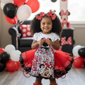 K&ouml;nnte beinhalten: Ein l&auml;chelndes Kind tr&auml;gt ein Outfit zum Thema Minnie Mouse. Das Outfit umfasst ein wei&szlig;es Oberteil, einen roten T&uuml;llrock mit schwarzen Punkten und einen passenden Kordelzugbeutel. Rote Schleifen schm&uuml;cken das Haar des Kindes. Ballons in Rot, Wei&szlig; und Schwarz sind im Hintergrund.