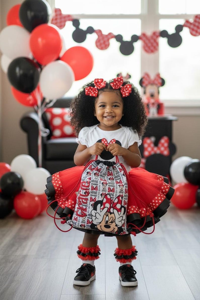 K&ouml;nnte beinhalten: Ein l&auml;chelndes Kind tr&auml;gt ein Outfit zum Thema Minnie Mouse. Das Outfit umfasst ein wei&szlig;es Oberteil, einen roten T&uuml;llrock mit schwarzen Punkten und einen passenden Kordelzugbeutel. Rote Schleifen schm&uuml;cken das Haar des Kindes. Ballons in Rot, Wei&szlig; und Schwarz sind im Hintergrund.