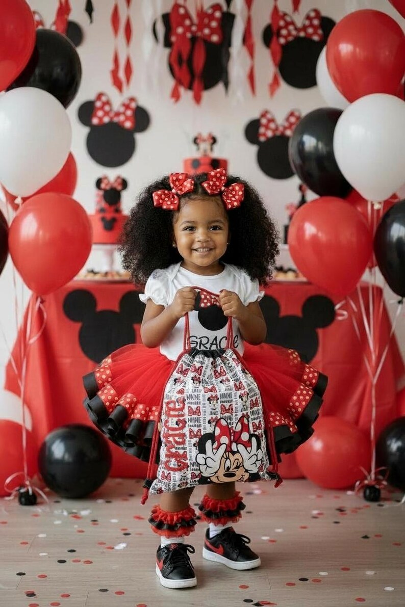 K&ouml;nnte beinhalten: Ein l&auml;chelndes Kind h&auml;lt einen Minnie Mouse-Kordelzugbeutel. Sie tr&auml;gt einen rot-schwarzen T&uuml;llrock, ein wei&szlig;es Oberteil und schwarze Turnschuhe. Rote, wei&szlig;e und schwarze Luftballons und Dekorationen schaffen eine festliche Atmosph&auml;re.