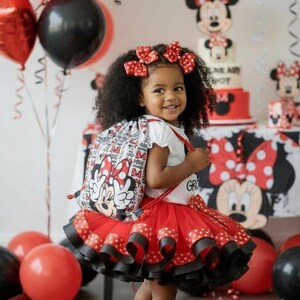 K&ouml;nnte beinhalten: Ein Kind in einem Minnie-Maus-Outfit, mit einem roten Tutu-Rock, Rucksack und Haarschleifen. Der Hintergrund umfasst rote und schwarze Luftballons, eine Torte und Dekorationen, was eine festliche Atmosph&auml;re schafft, wahrscheinlich eine Geburtstagsfeier.
