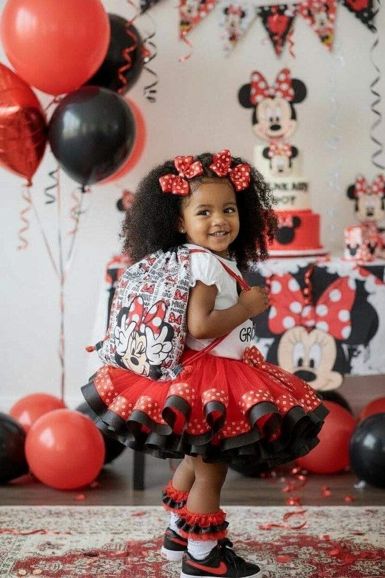 K&ouml;nnte beinhalten: Ein Kind in einem Minnie-Maus-Outfit, mit einem roten Tutu-Rock, Rucksack und Haarschleifen. Der Hintergrund umfasst rote und schwarze Luftballons, eine Torte und Dekorationen, was eine festliche Atmosph&auml;re schafft, wahrscheinlich eine Geburtstagsfeier.