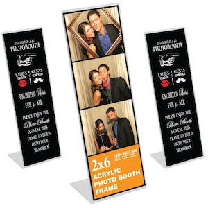 May include: A clear acrylic photo booth frame with a black and white design. The frame is 2x6 inches and has a slot for a photo strip. The frame is on a white background. The text on the frame says "Step Right Up to the Photo Booth" and "Unlimited Photos Fun for All".