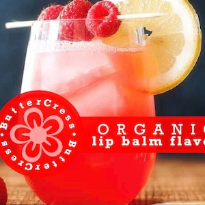 May include: A glass of pink lemonade with raspberries and a lemon wedge. The glass is on a wooden surface. The text "ORGANIC lip balm flavor" is on a red banner.