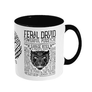 May include: Black and white mug with a black interior. The mug features a graphic of a wolf with the text "Feral Druid Powerful Pussycat Savage Bites & Powerful Roars Killer Kitty Strikes Again Bitten By A Kitten Tis But A Scratch Try To Hide The Scent Of Blood & Find Yourself Beneath The Mud".