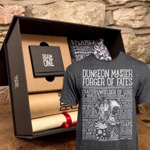 May include: A gift box set for a Dungeon Master, featuring a grey t-shirt with the text "Dungeon Master Forger of Fates" and a list of duties, a brown paper scroll, a small black box with the text "Realm One", and a black bandana with the text "Eflame & the occasional gamble don't..."