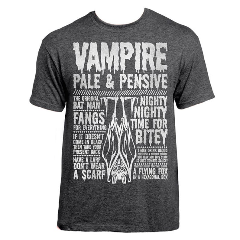 May include: A dark gray t-shirt with a white graphic print that reads "Vampire Pale & Pensive" and includes a bat illustration. The text includes the phrases "The Original Bat Man", "Fangs for Everything", "Nighty Nighty Time for Bitey", "Have a Larf Don't Wear a Scarf", and "A Flying Fox in a Hexagonal Box".