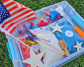 May include: A clear plastic storage bin filled with red, white, and blue sensory items for a patriotic themed activity. The bin contains red and blue rice, wooden stars, red, white, and blue pom poms, and a small American flag.