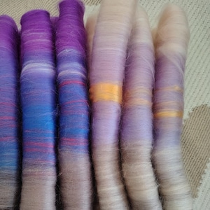 Op de afbeelding: Een verzameling handgeverfde garenklossen in tinten paars, blauw en beige. Het garen is gerangschikt in een stapel, met de paarse en blauwe klossen bovenop en de beige klossen onderaan.