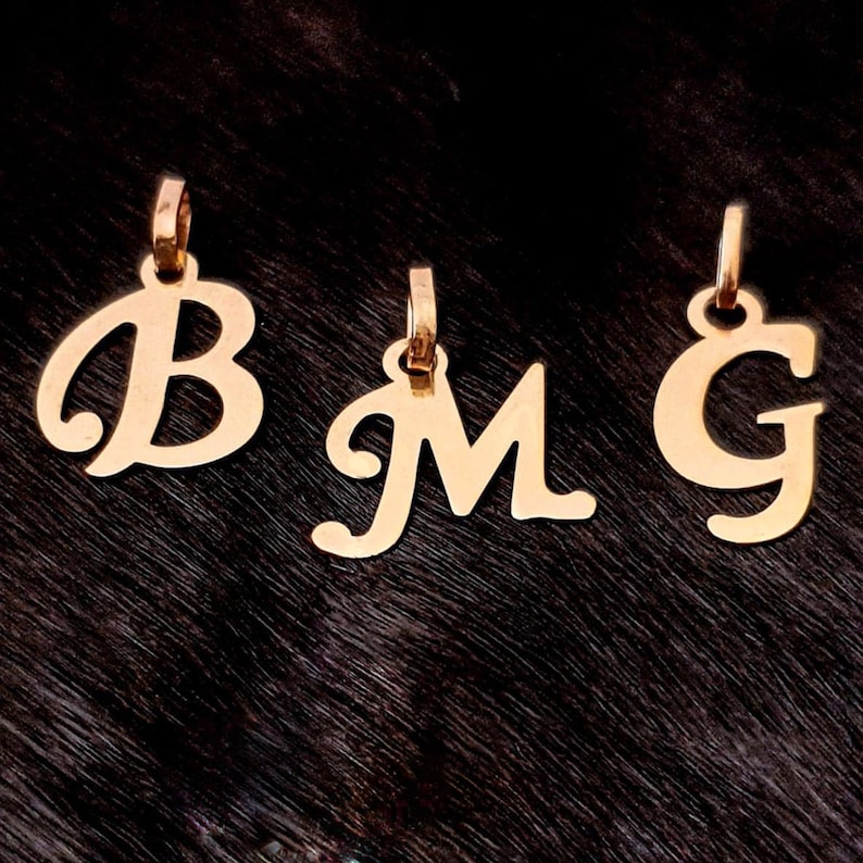 Puede incluir: Tres colgantes de metal en tono dorado con las letras B, M y G en una fuente cursiva. Cada colgante incluye una peque&ntilde;a anilla circular en la parte superior para sujetarlo a una cadena, mostrando un acabado pulido y formas de letra claras.