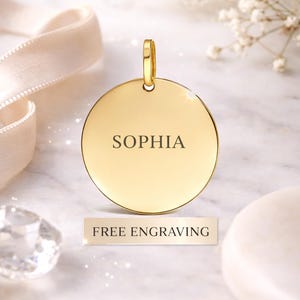 May include: A gold-coloured, circular pendant engraved with the name "SOPHIA". The pendant has a loop for a chain. A small rectangular sign below reads "FREE ENGRAVING". The background features a ribbon and flowers, with a marble effect.