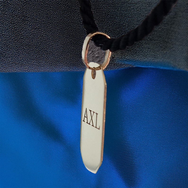 May include: A silver tag with the word "AXL" engraved on it hangs from a black cord. The tag is shaped like a long, narrow oval with rounded edges.