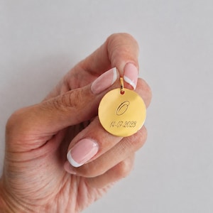 Puede incluir: Un colgante redondo dorado con un peque&ntilde;o lazo en la parte superior. El colgante presenta una letra cursiva "C" grabada y la fecha "14-07-2023". El colgante es sostenido por una mano con esmalte de u&ntilde;as rosa.