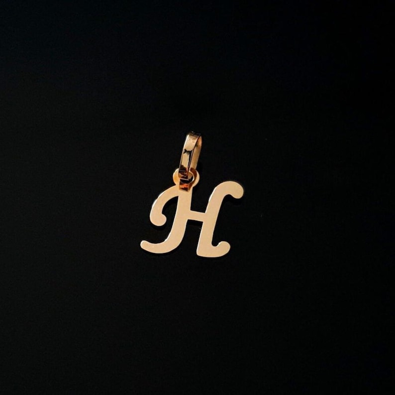 Puede incluir: Un colgante de metal en tono dorado con forma de letra may&uacute;scula H en una fuente cursiva. El colgante presenta una superficie pulida y reflectante e incluye una peque&ntilde;a anilla circular integrada en la parte superior para una cadena.
